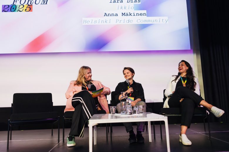 Kolme henkilöä osallistumassa Helsinki Pride Business Forumin paneelikeskusteluun, osallistujat näyttävät iloisilta ja istuvat lavalla powerpoint-esityksen edessä